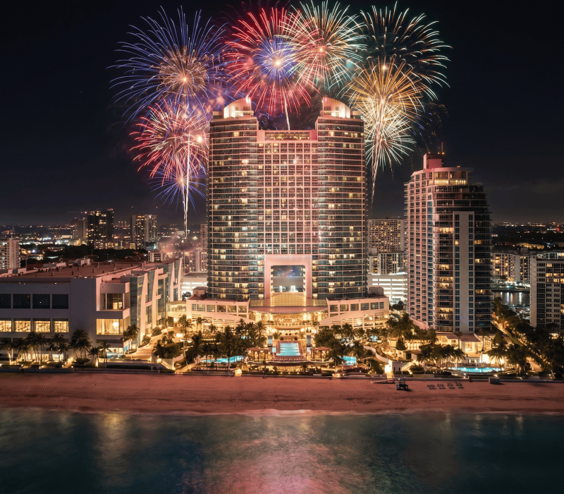 Colorful fireworks burst above a large, illuminated hotel complex on a beach, with city lights and the ocean at night.