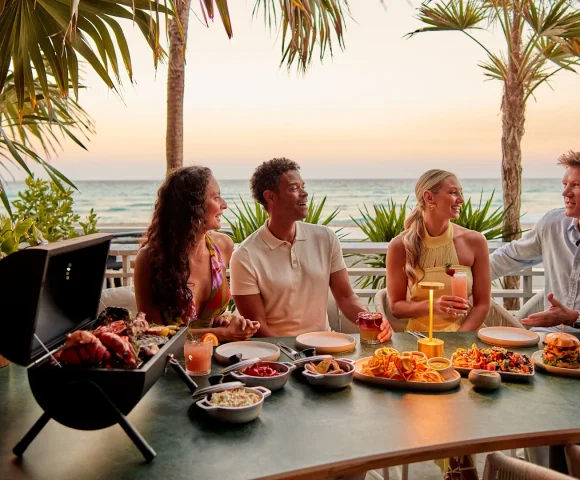 Four friends dining outdoors at a beachside resort table with a small tabletop grill and sunset ocean views.