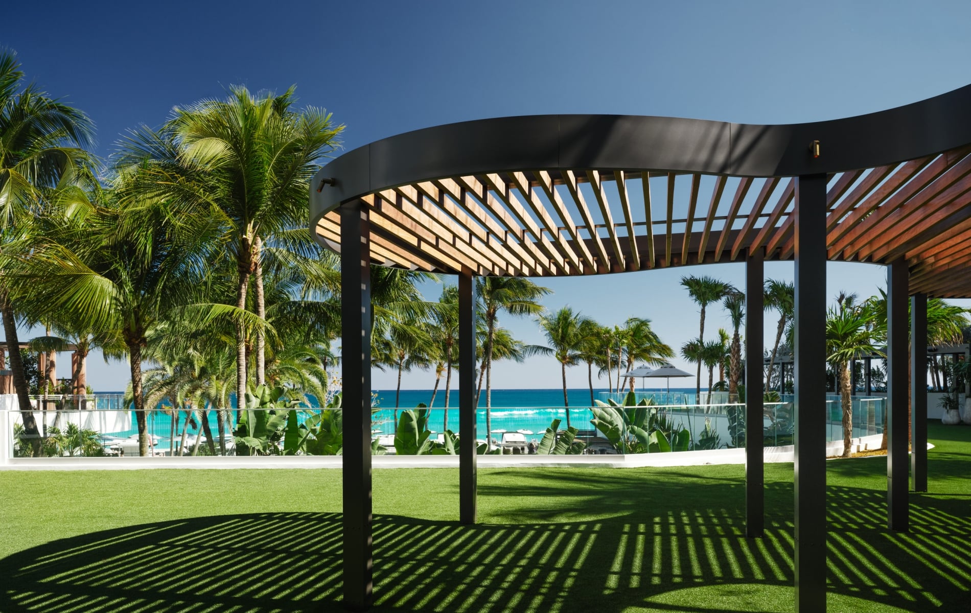 A curved black and wood slatted pergola casts striped shadows on a green lawn, with palm trees and a turquoise ocean visible in the background under a clear sky.