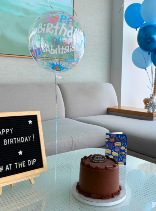 A chocolate birthday cake, a letter board, and blue balloons are arranged on a glass table. The board reads: "HAPPY BIRTHDAY! # AT THE DIP" in front of a grey sofa.