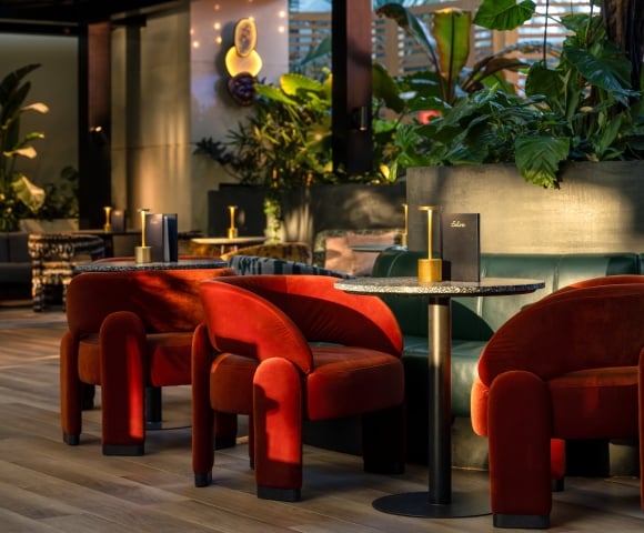 An elegant modern lounge featuring plush rust-orange velvet armchairs, a small round marble-topped table with gold detailing, and lush indoor tropical plants in the background.