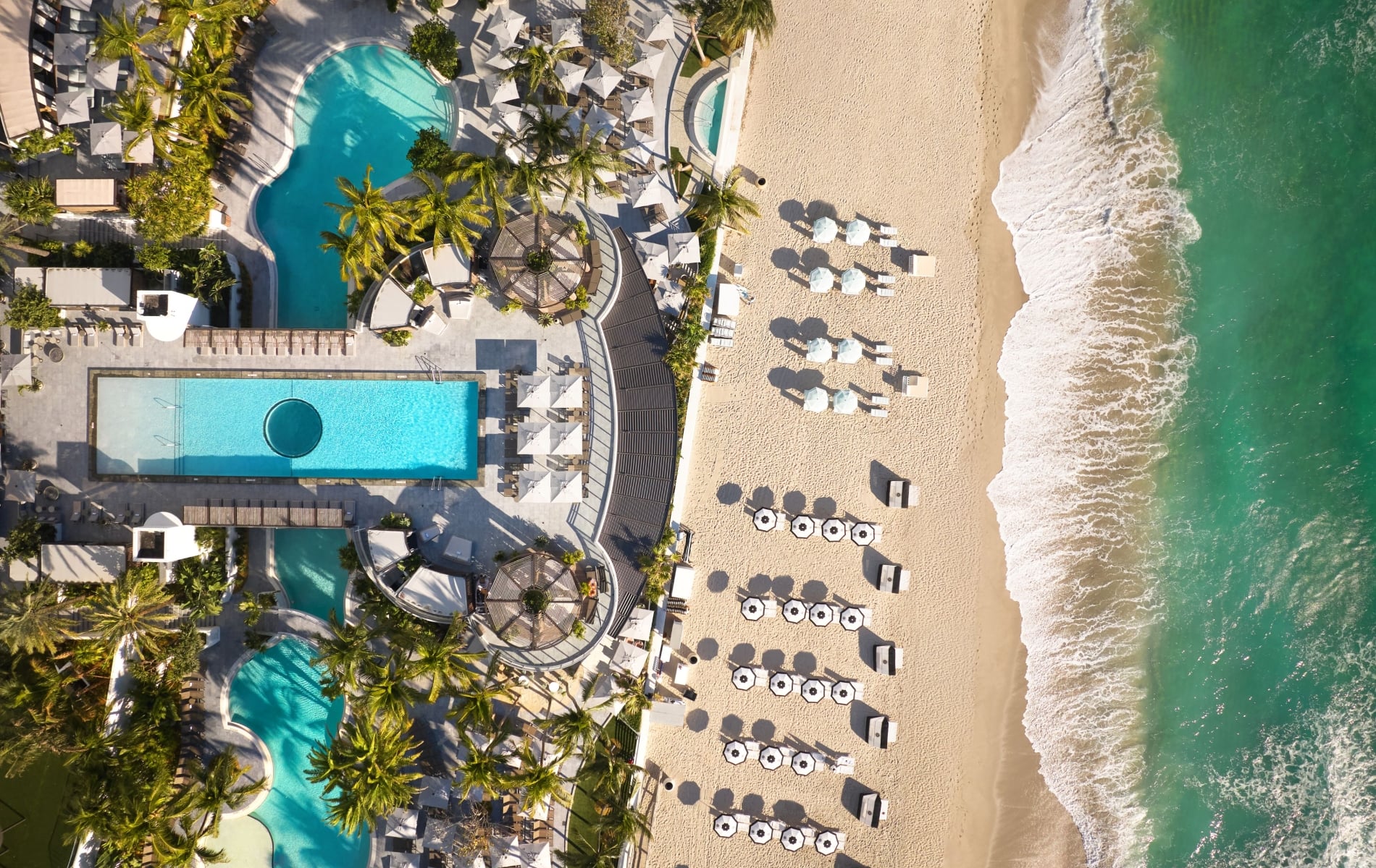 An aerial view of a beachfront resort pool, palm trees, and rows of umbrellas on the sand next to ocean waves.