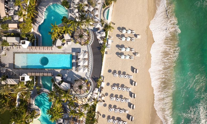 An aerial view of a beachfront resort pool, palm trees, and rows of umbrellas on the sand next to ocean waves.
