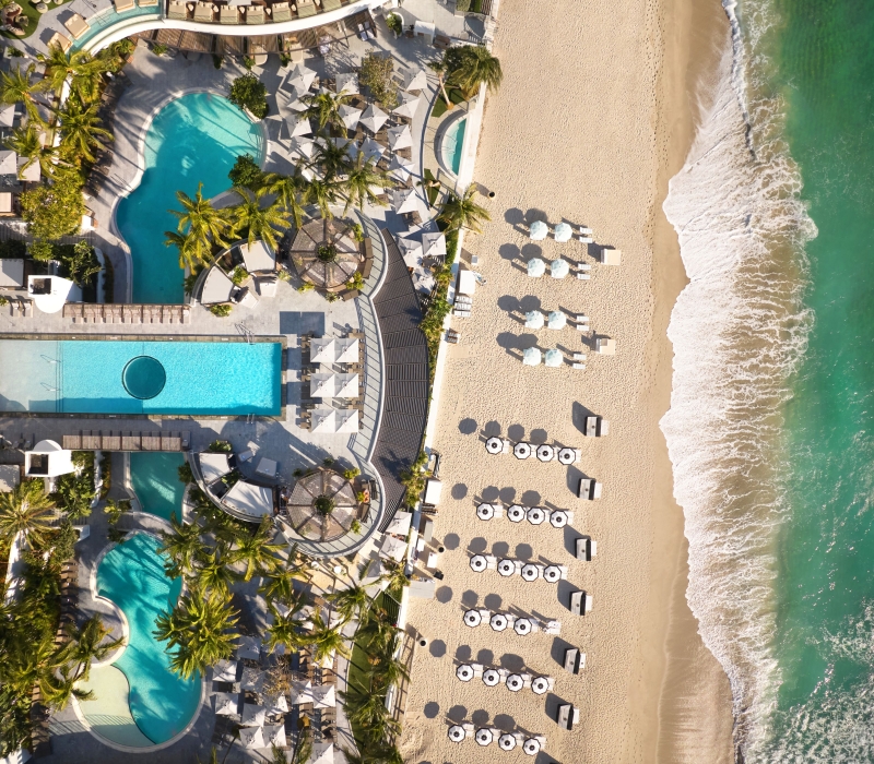 An aerial view of a beachfront resort pool, palm trees, and rows of umbrellas on the sand next to ocean waves.