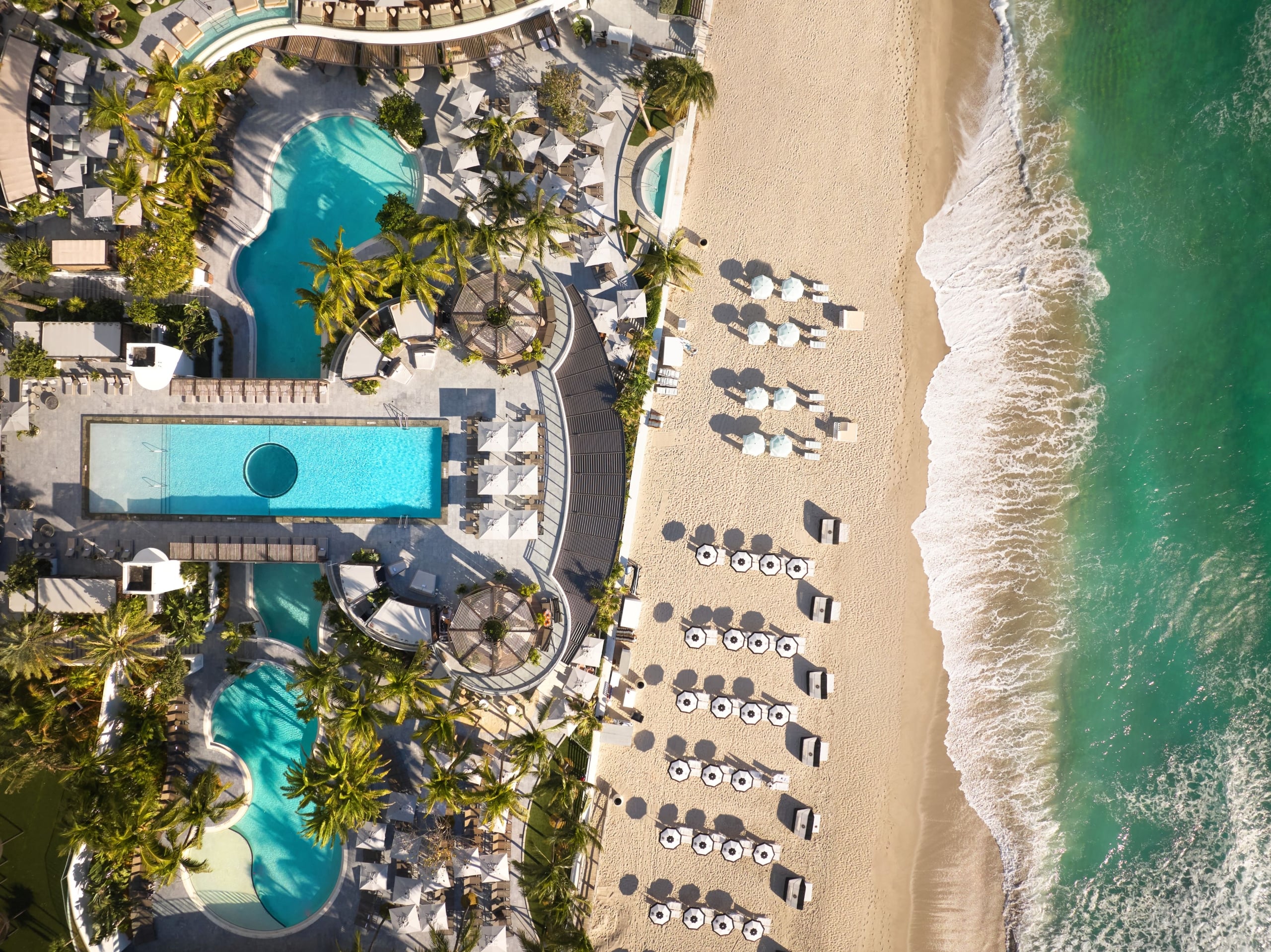 An aerial view of a beachfront resort pool, palm trees, and rows of umbrellas on the sand next to ocean waves.