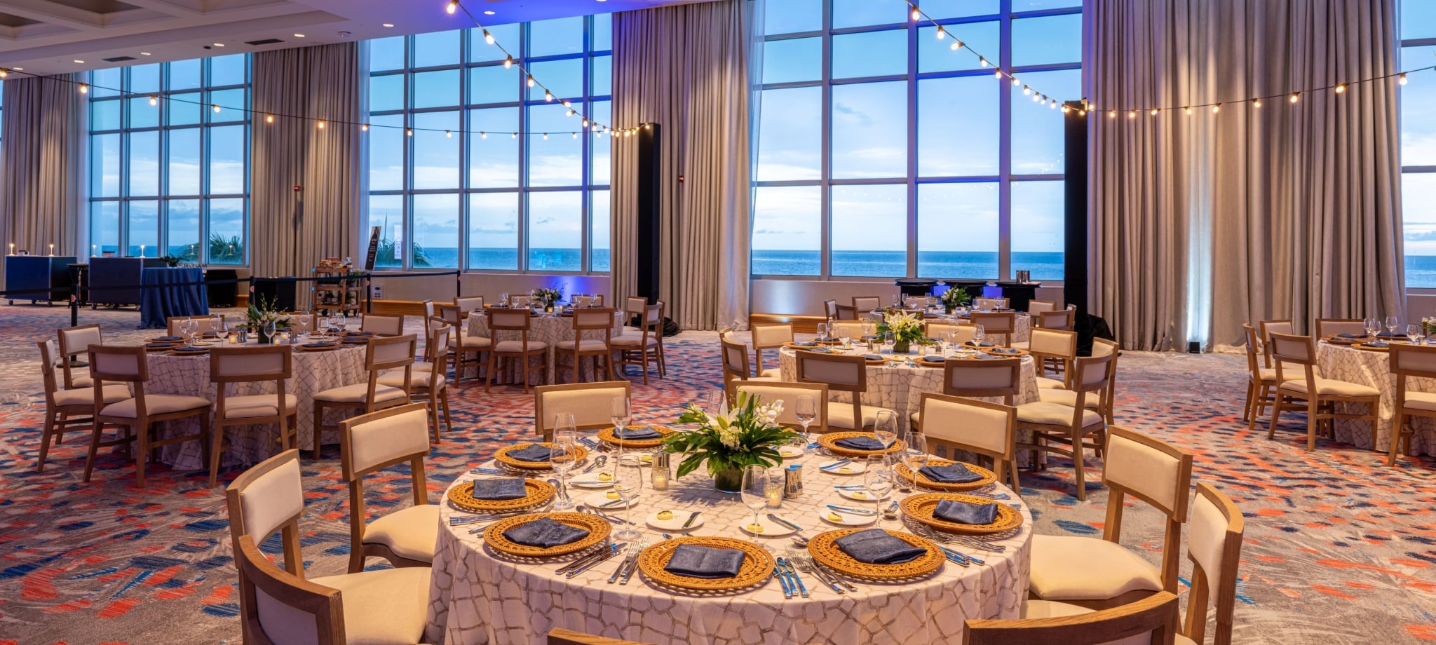 A large resort banquet room featuring round dining tables set with white linens, gold chargers, and dark blue napkins, illuminated by warm string lights under a high ceiling with tall windows overlooking the ocean.