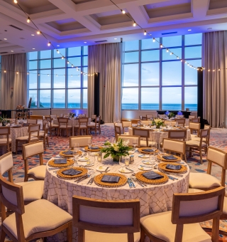 A large resort banquet room featuring round dining tables set with white linens, gold chargers, and dark blue napkins, illuminated by warm string lights under a high ceiling with tall windows overlooking the ocean.
