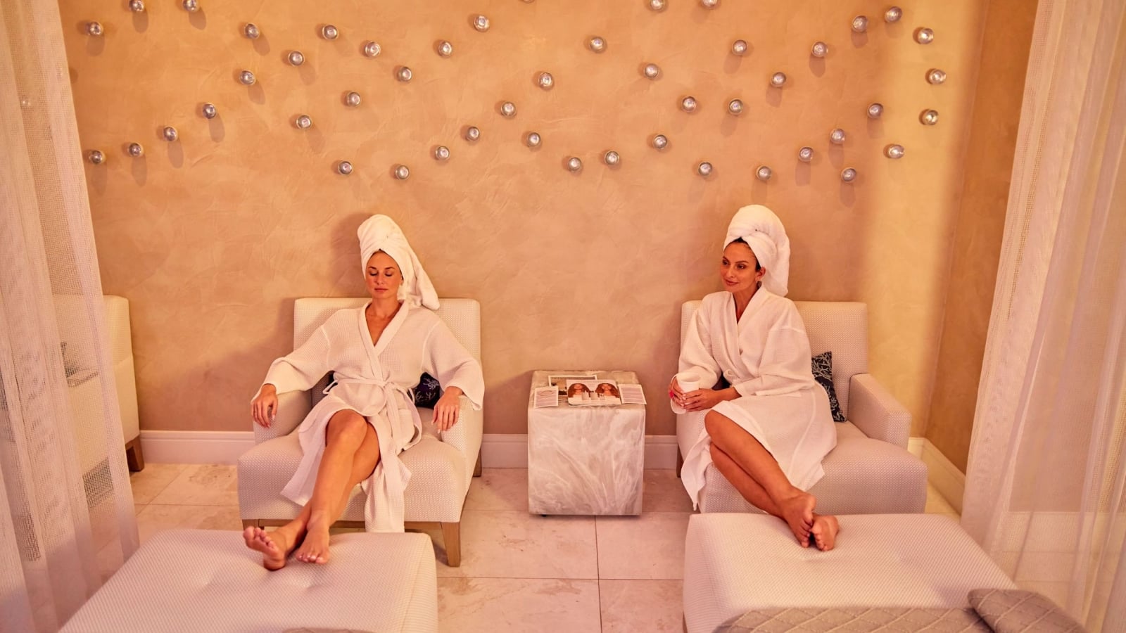 Two women in white robes and hair towels relax in lounge chairs inside a resort spa relaxation room.