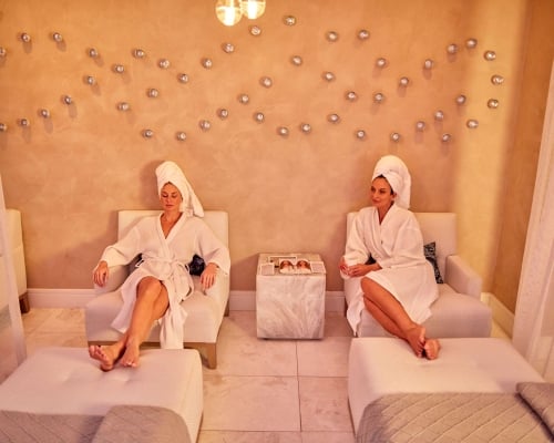 Two women in white robes and hair towels relax in lounge chairs inside a resort spa relaxation room.