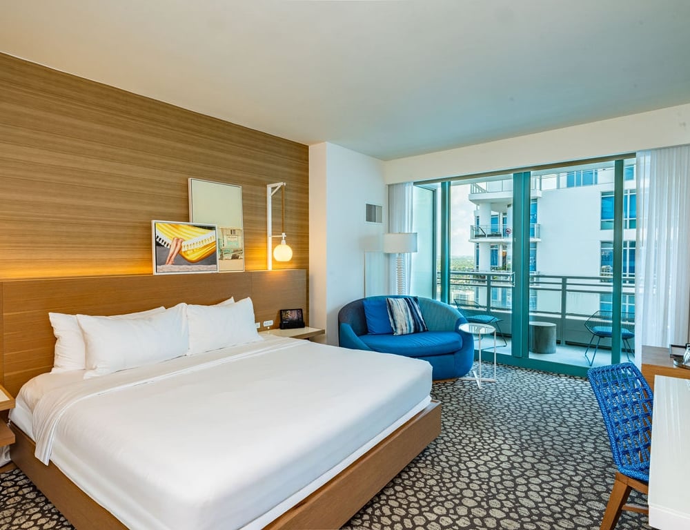 A modern hotel room features a large bed, a blue circular couch, and a desk, all on patterned carpet, with a balcony offering a city view.