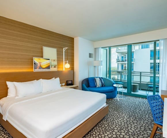 A modern hotel room features a large bed, a blue circular couch, and a desk, all on patterned carpet, with a balcony offering a city view.