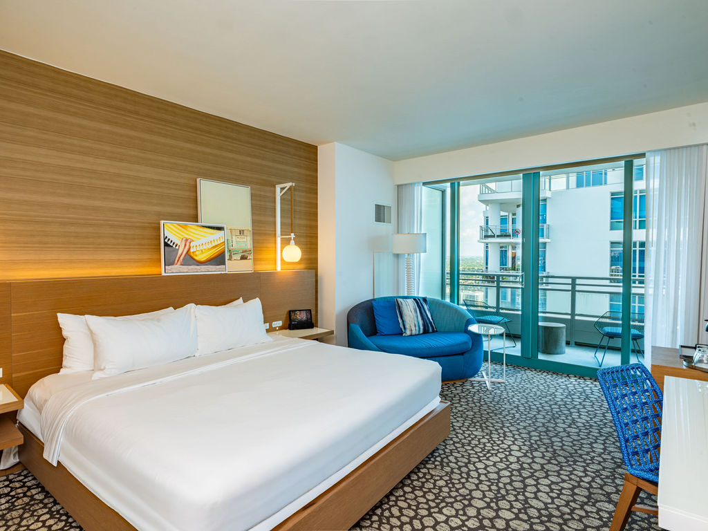 A modern hotel room features a large bed, a blue circular couch, and a desk, all on patterned carpet, with a balcony offering a city view.