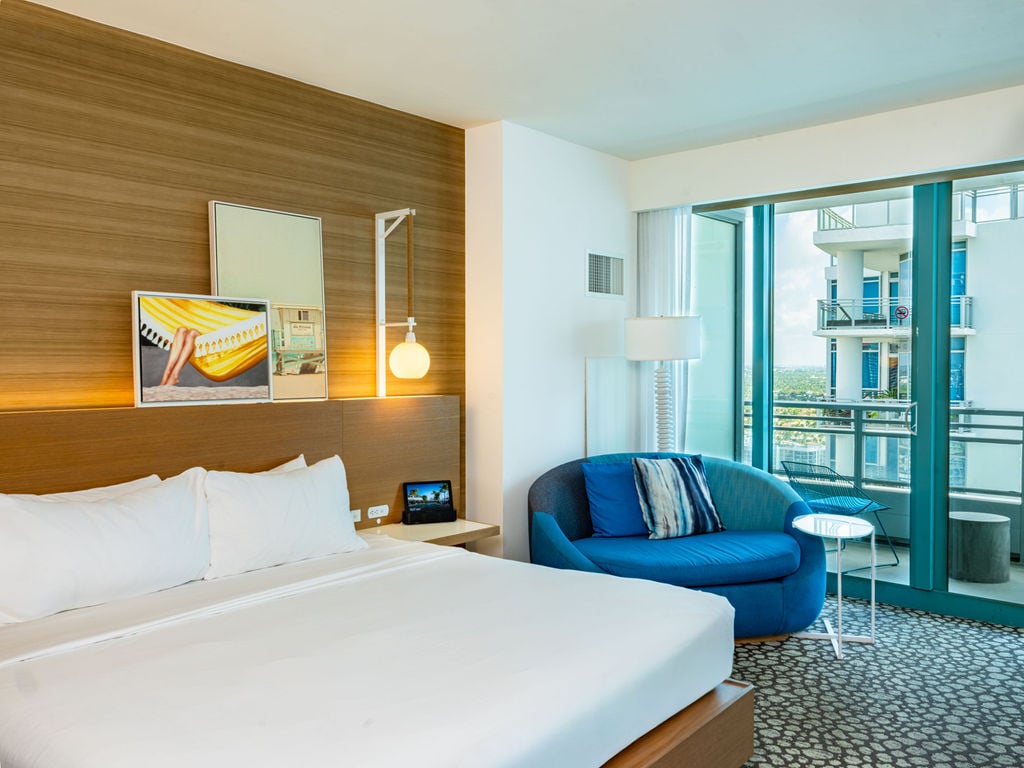 Modern bedroom with a white bed, blue lounge chair, and wall art. Glass doors open to a balcony with a city view, overlooking another building.