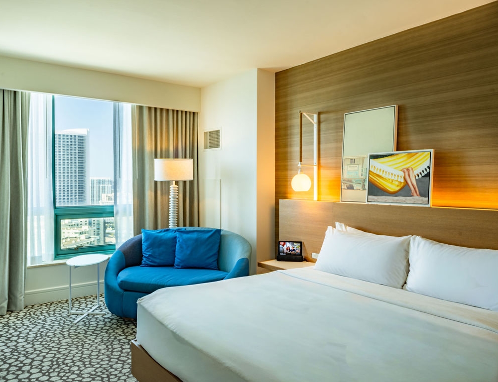 A modern hotel room features a large white bed, a blue chaise lounge by a city-view window, and a wooden accent wall with contemporary art.
