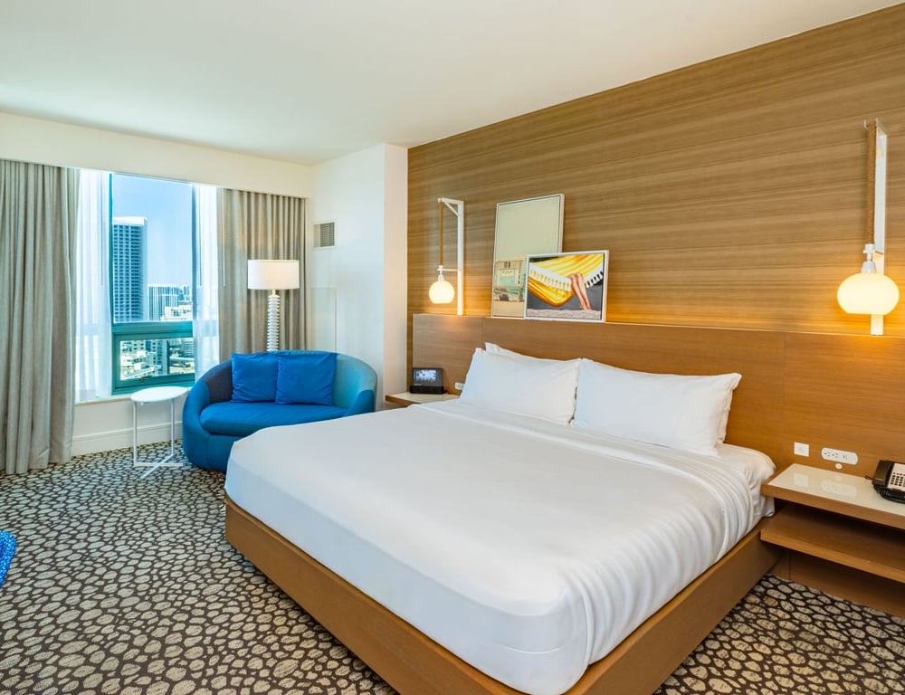 A modern hotel bedroom is furnished with a king-sized bed, a blue loveseat, and a city-view window. The room features a wood-paneled accent wall, patterned carpet, and bedside lighting.