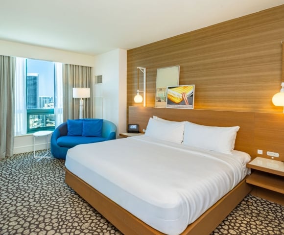 A modern hotel bedroom is furnished with a king-sized bed, a blue loveseat, and a city-view window. The room features a wood-paneled accent wall, patterned carpet, and bedside lighting.