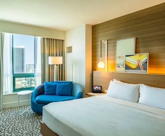 A modern hotel room features a large white bed, a blue chaise lounge by a city-view window, and a wooden accent wall with contemporary art.
