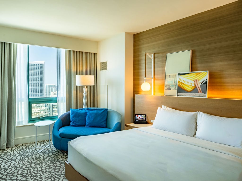 A modern hotel room features a large white bed, a blue chaise lounge by a city-view window, and a wooden accent wall with contemporary art.