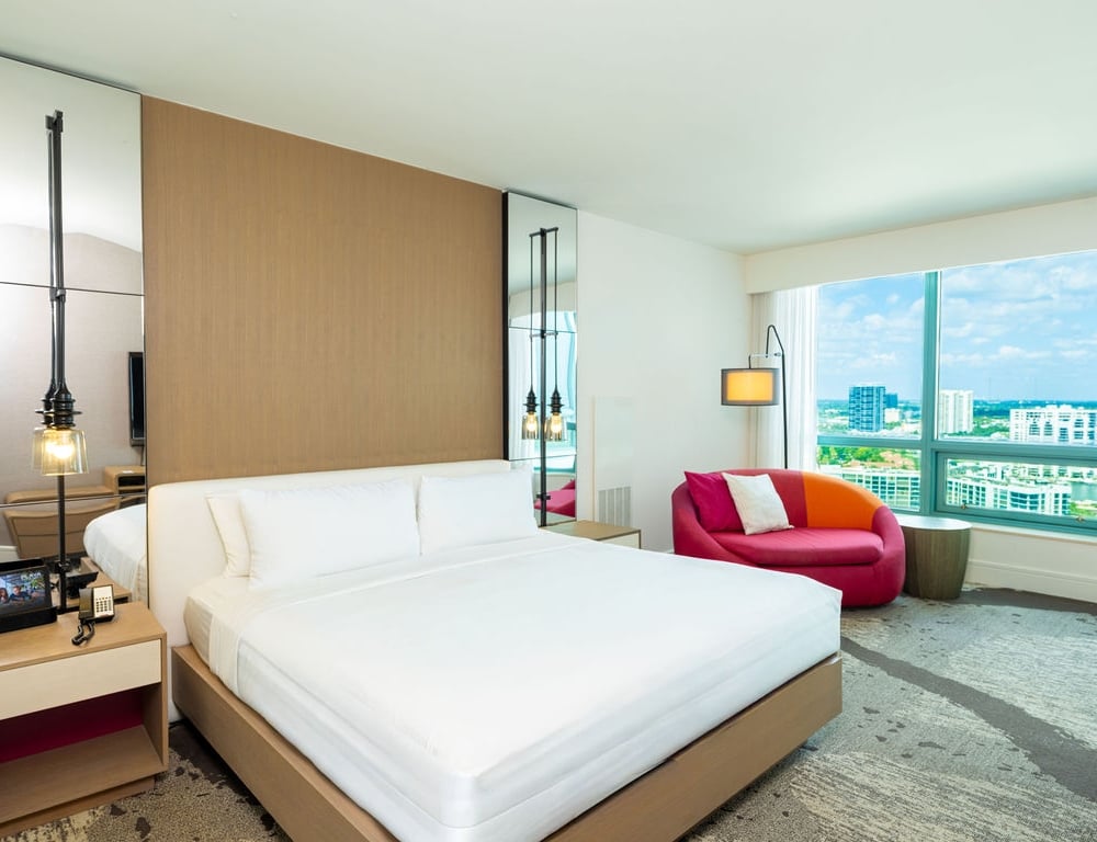 A modern hotel bedroom features a large white bed and a colorful pink/orange chair, with a screen displaying "PLAZA", overlooking a city skyline through large windows.