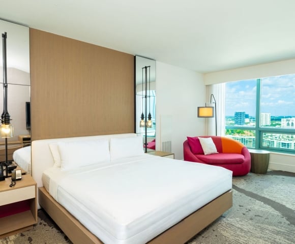 A modern hotel bedroom features a large white bed and a colorful pink/orange chair, with a screen displaying "PLAZA", overlooking a city skyline through large windows.