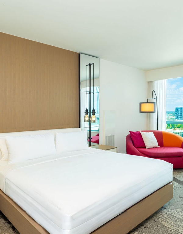 A modern hotel bedroom features a large white bed and a colorful pink/orange chair, with a screen displaying "PLAZA", overlooking a city skyline through large windows.