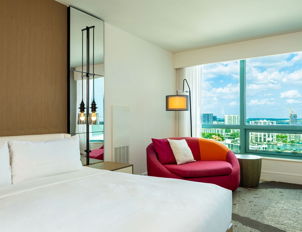 A neatly made bed with white linens is in a modern hotel room, featuring a colorful sofa, floor lamp, and a large window overlooking a city skyline and water.