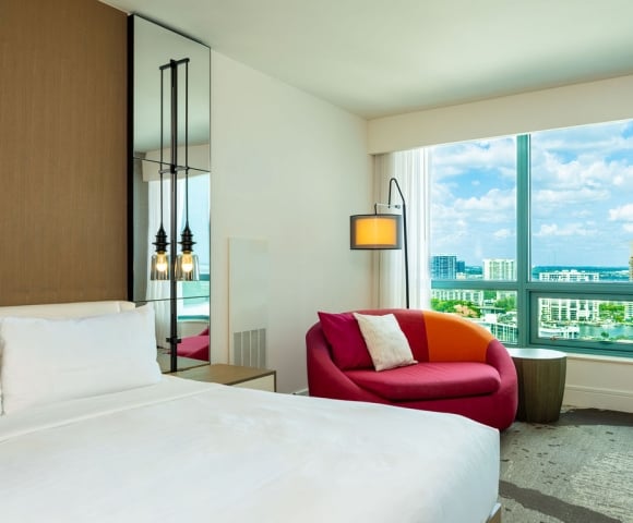 A neatly made bed with white linens is in a modern hotel room, featuring a colorful sofa, floor lamp, and a large window overlooking a city skyline and water.