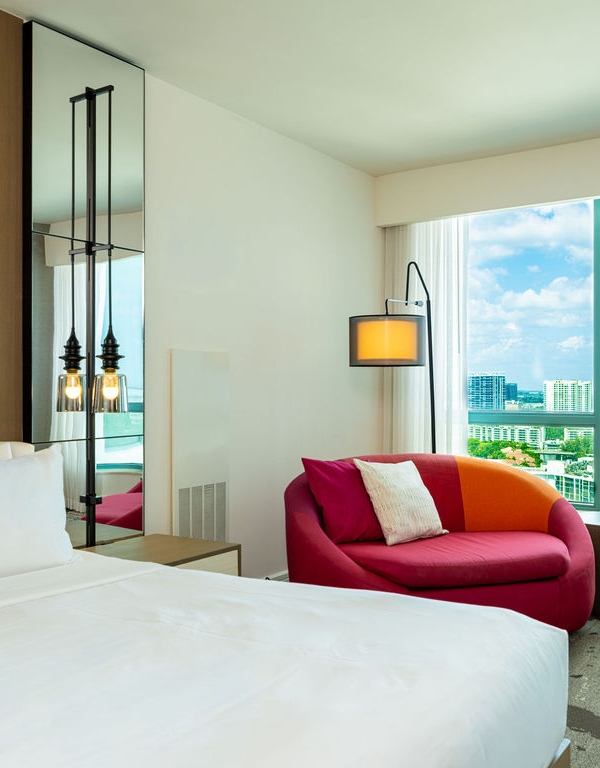 A neatly made bed with white linens is in a modern hotel room, featuring a colorful sofa, floor lamp, and a large window overlooking a city skyline and water.