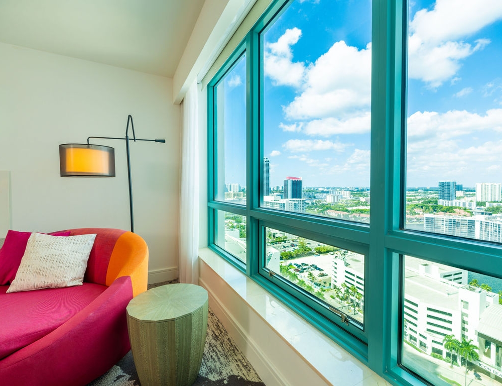 A pink and orange chaise lounge sits beside a large window overlooking a city skyline under a blue, cloudy sky. Text visible: ICON BEACH.