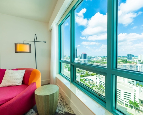 A pink and orange chaise lounge sits beside a large window overlooking a city skyline under a blue, cloudy sky. Text visible: ICON BEACH.