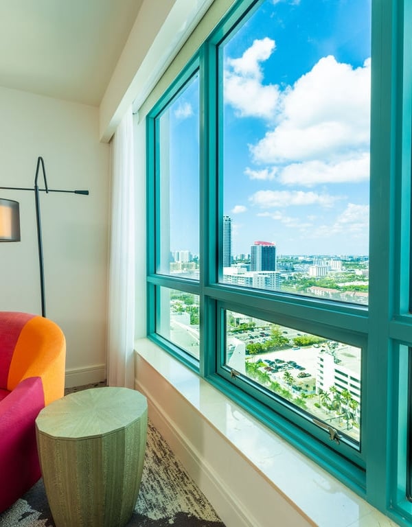 A pink and orange chaise lounge sits beside a large window overlooking a city skyline under a blue, cloudy sky. Text visible: ICON BEACH.
