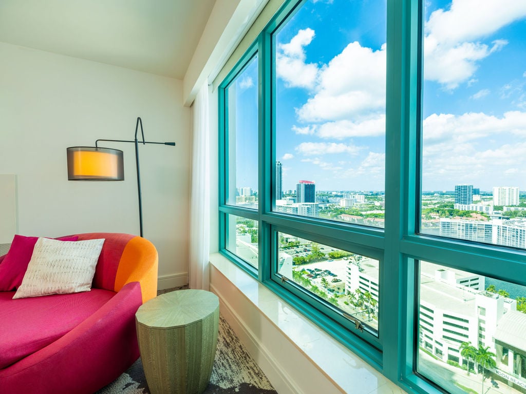A pink and orange chaise lounge sits beside a large window overlooking a city skyline under a blue, cloudy sky. Text visible: ICON BEACH.