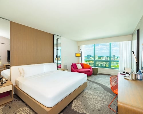 Brightly lit, modern hotel room featuring a king bed, red sofa, and desk. A large window offers expansive views of a city and water.