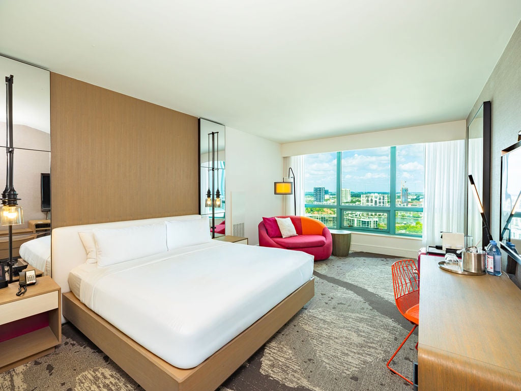 Brightly lit, modern hotel room featuring a king bed, red sofa, and desk. A large window offers expansive views of a city and water.