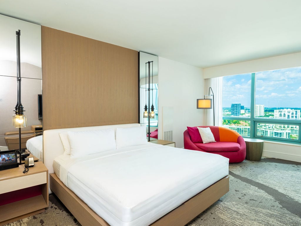 A modern hotel bedroom features a large white bed and a colorful pink/orange chair, with a screen displaying "PLAZA", overlooking a city skyline through large windows.