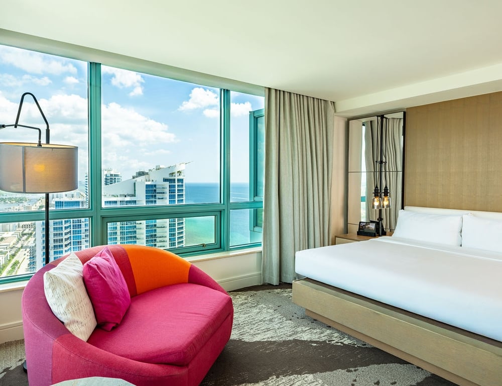 A luxurious hotel bedroom is furnished with a large bed and a vibrant pink armchair, overlooking a city skyline and ocean through floor-to-ceiling windows.