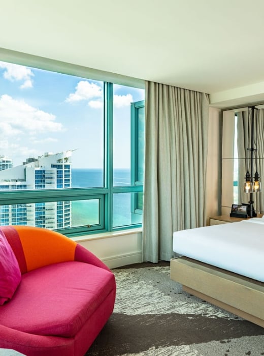 A luxurious hotel bedroom is furnished with a large bed and a vibrant pink armchair, overlooking a city skyline and ocean through floor-to-ceiling windows.