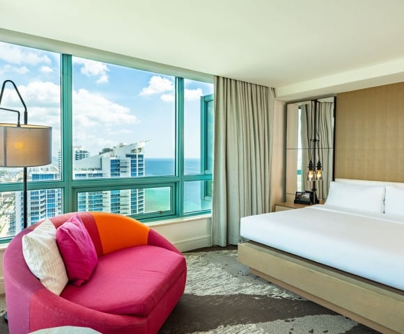 A luxurious hotel bedroom is furnished with a large bed and a vibrant pink armchair, overlooking a city skyline and ocean through floor-to-ceiling windows.
