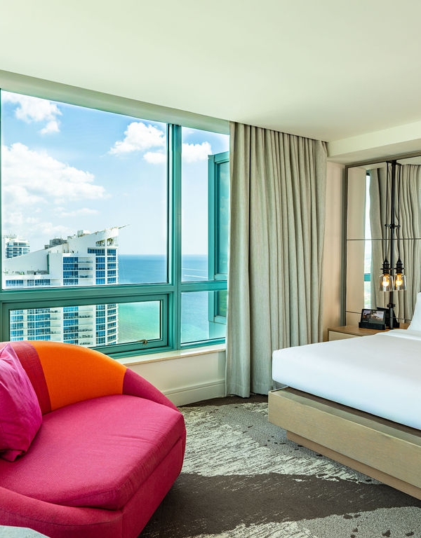 A luxurious hotel bedroom is furnished with a large bed and a vibrant pink armchair, overlooking a city skyline and ocean through floor-to-ceiling windows.