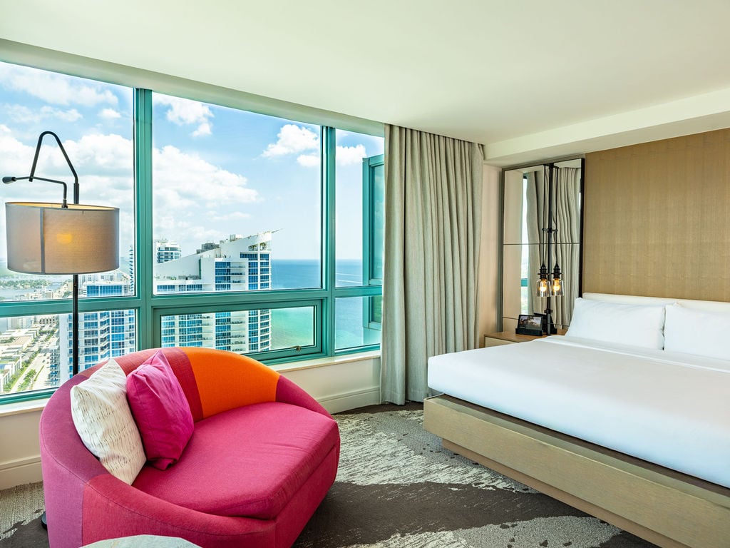 A luxurious hotel bedroom is furnished with a large bed and a vibrant pink armchair, overlooking a city skyline and ocean through floor-to-ceiling windows.