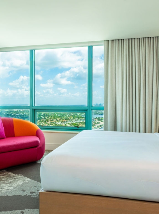 A modern hotel bedroom features a white bed and a vibrant pink sofa, positioned by large windows offering a high-rise view of a city and bay under a cloudy sky.