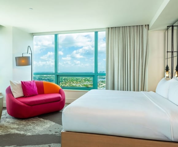 A modern hotel bedroom features a white bed and a vibrant pink sofa, positioned by large windows offering a high-rise view of a city and bay under a cloudy sky.