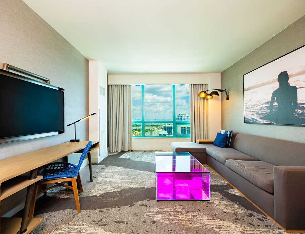A modern hotel room features an L-shaped sofa, TV, and desk, with a city/ocean view. A clear coffee table glows pink, resting on a patterned carpet.