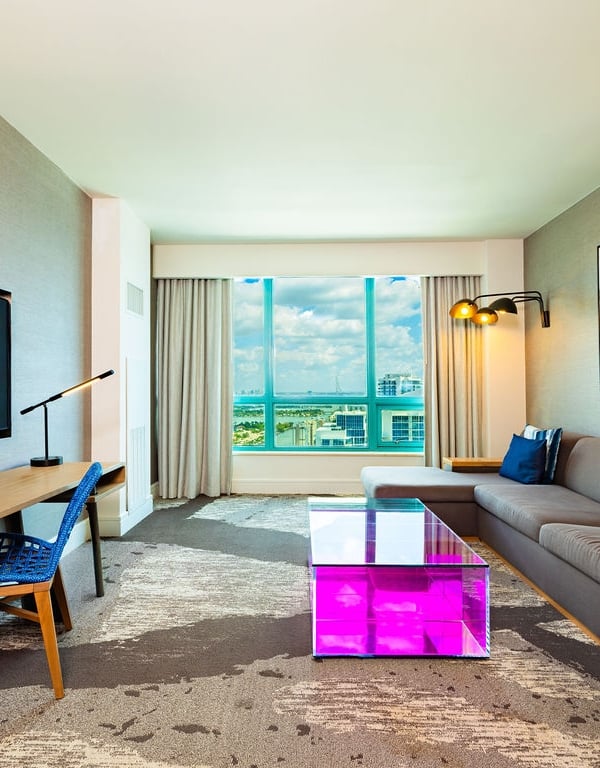 A modern hotel room features an L-shaped sofa, TV, and desk, with a city/ocean view. A clear coffee table glows pink, resting on a patterned carpet.