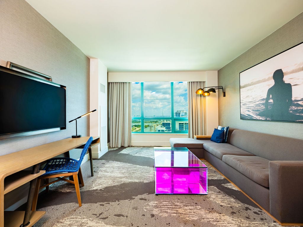 A modern hotel room features an L-shaped sofa, TV, and desk, with a city/ocean view. A clear coffee table glows pink, resting on a patterned carpet.