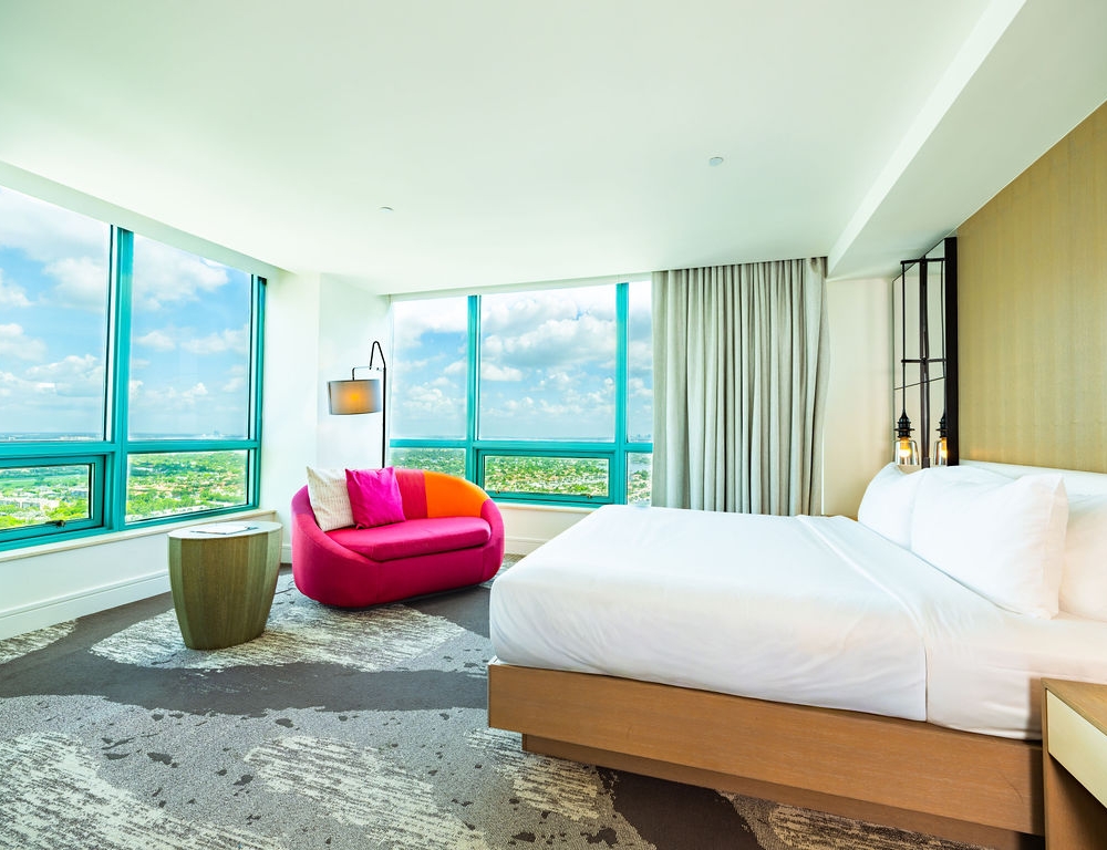 A modern hotel room features a white bed and a vibrant pink and orange sofa by expansive turquoise-framed windows, offering a scenic view of a city landscape.
