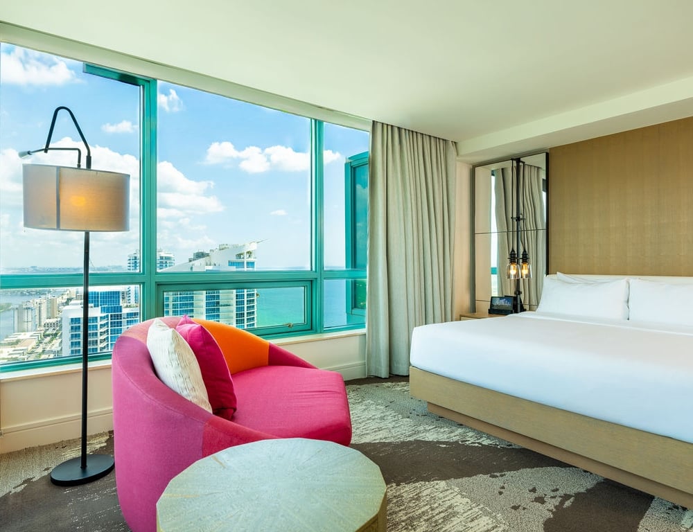 A modern hotel room offers a large bed and a bright pink armchair, overlooking a city skyline and ocean through floor-to-ceiling windows.