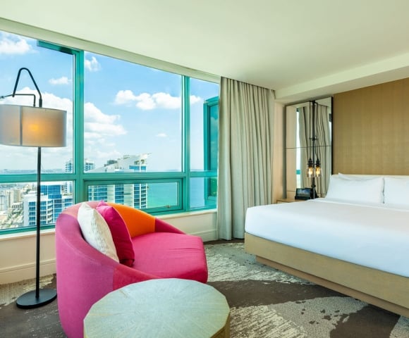 A modern hotel room offers a large bed and a bright pink armchair, overlooking a city skyline and ocean through floor-to-ceiling windows.