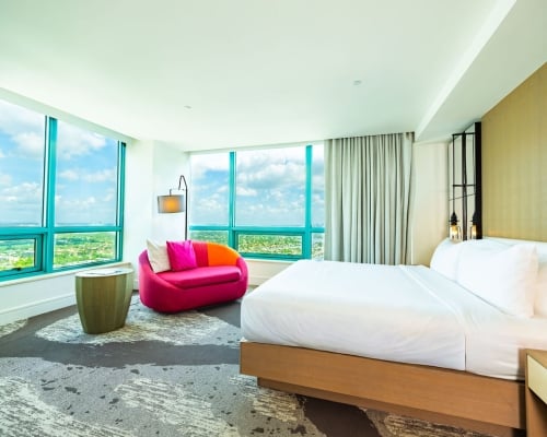 A modern hotel room features a white bed and a vibrant pink and orange sofa by expansive turquoise-framed windows, offering a scenic view of a city landscape.