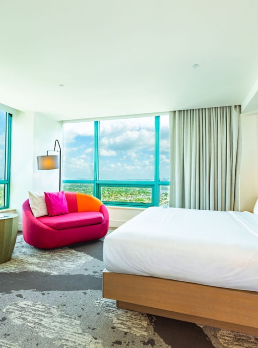 A modern hotel room features a white bed and a vibrant pink and orange sofa by expansive turquoise-framed windows, offering a scenic view of a city landscape.